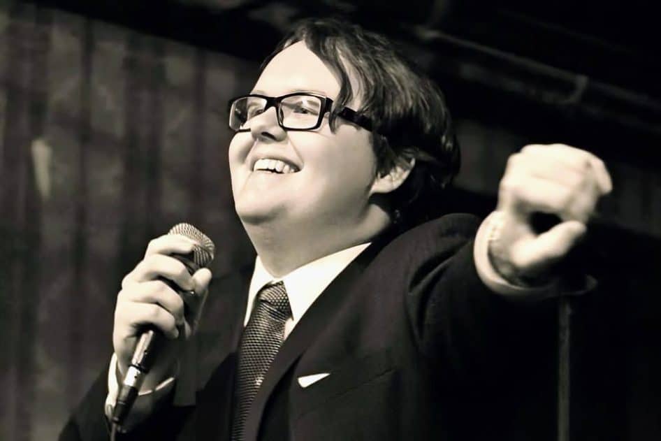 Connor (Mandy) Duff holding a microphone in right hand and extending his left hand to support the Connor Duff Memorial Bursary crowdfunding campaign.